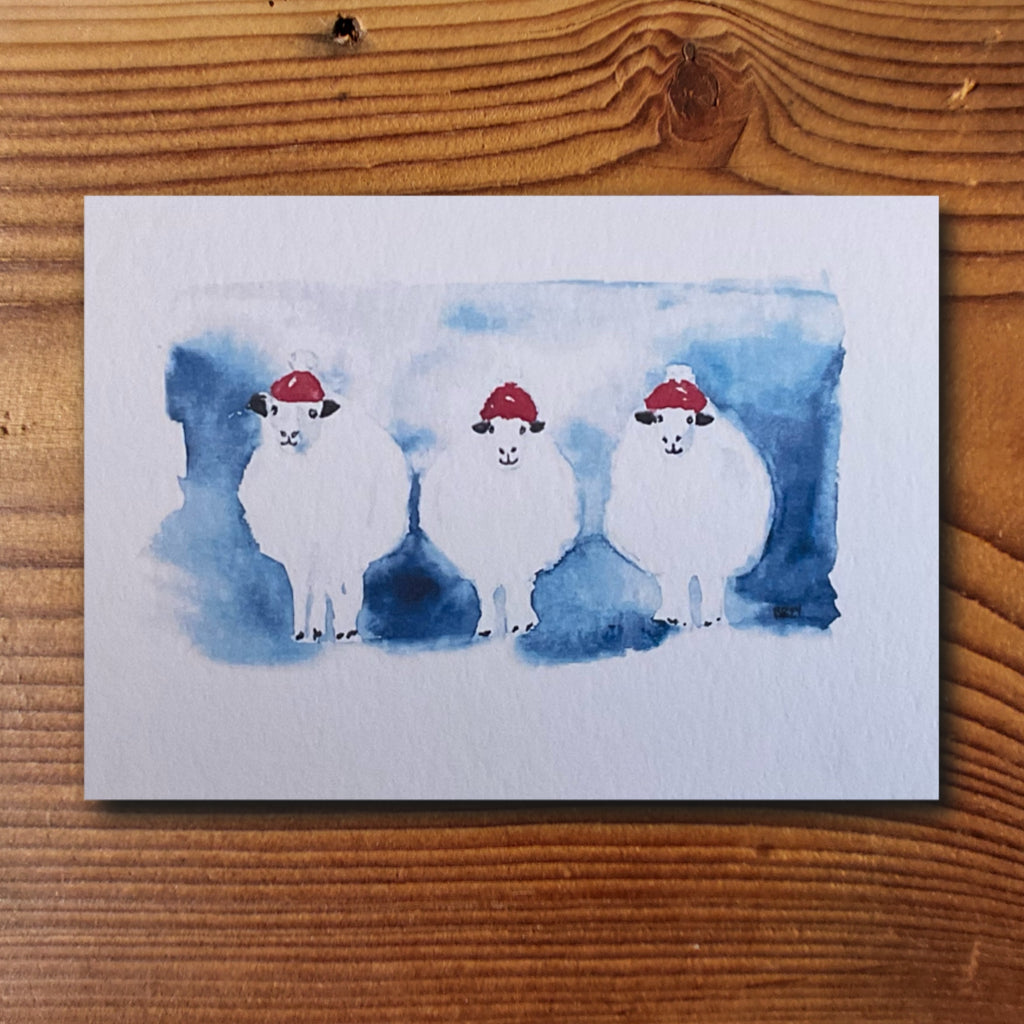 Weihnachtskarte mit drei Aquarell-Schafen in roten Weihnachtsmützen auf blauem Hintergrund