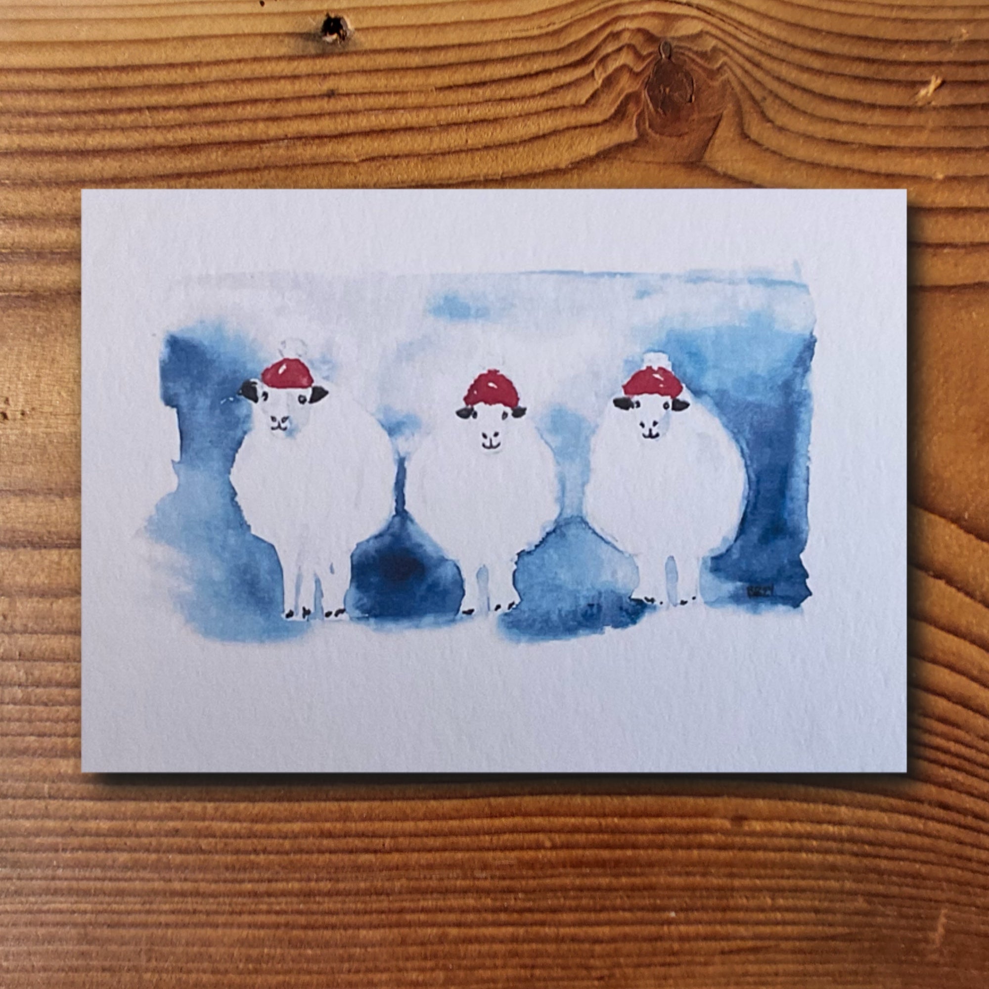 Weihnachtskarte mit drei Aquarell-Schafen in roten Weihnachtsmützen auf blauem Hintergrund
